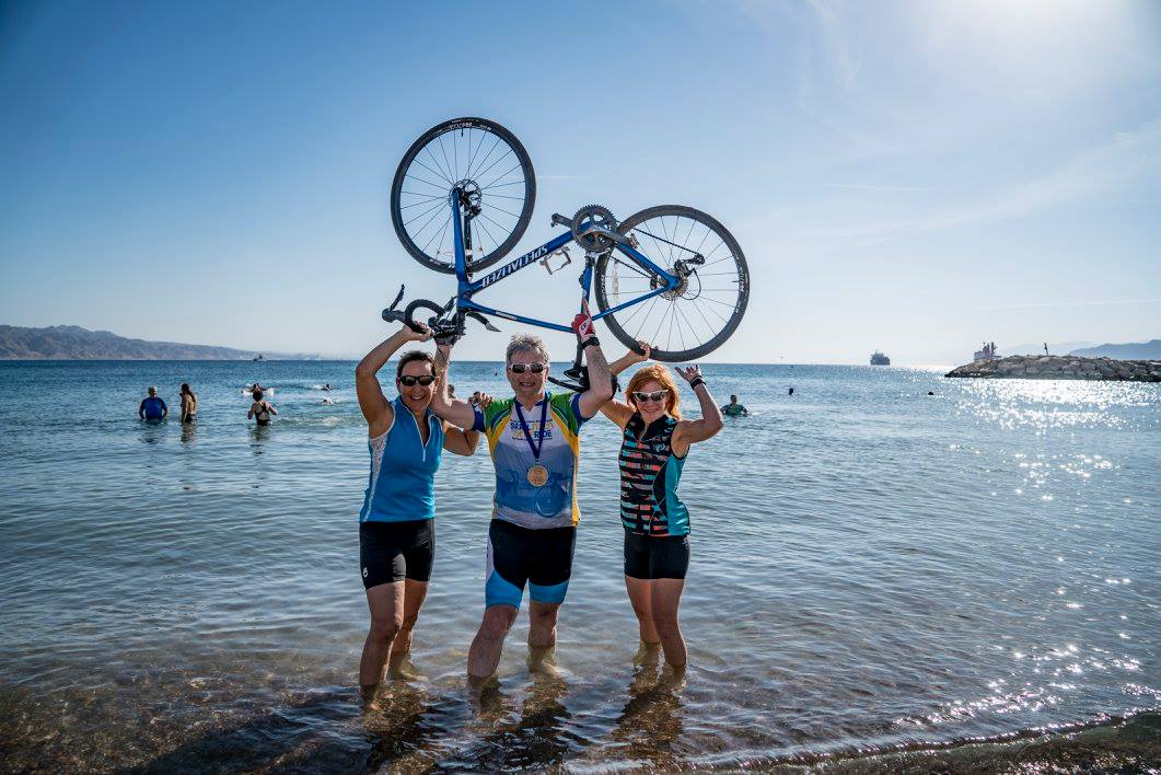 David Eisenberg in the water on the beach holding his bike in the air with two female riders